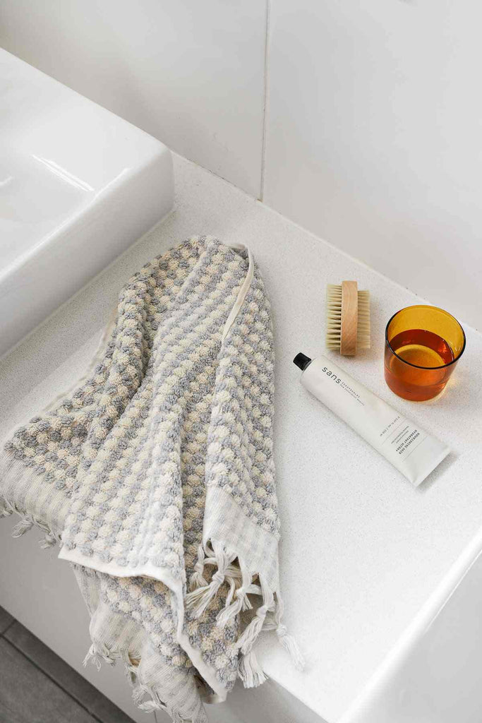Pale grey pompom Turkish cotton hand towel on white bathroom counter with brush and amber glass