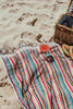Multicolour striped Turkish cotton bath towel on sandy beach with picnic basket and drink