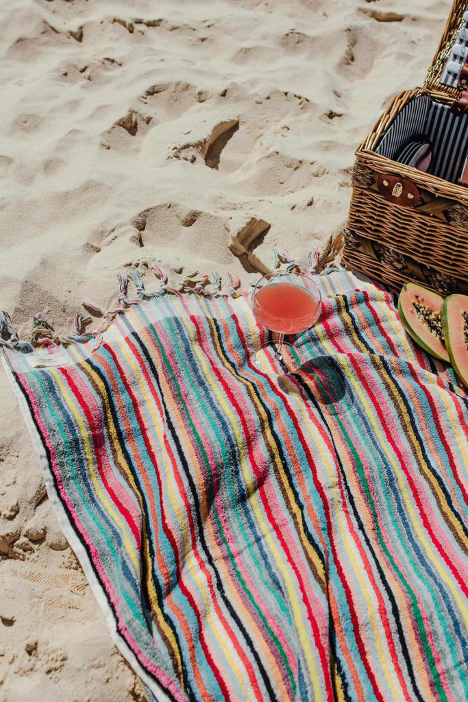 Multicolour striped Turkish cotton bath towel on sandy beach with picnic basket and drink